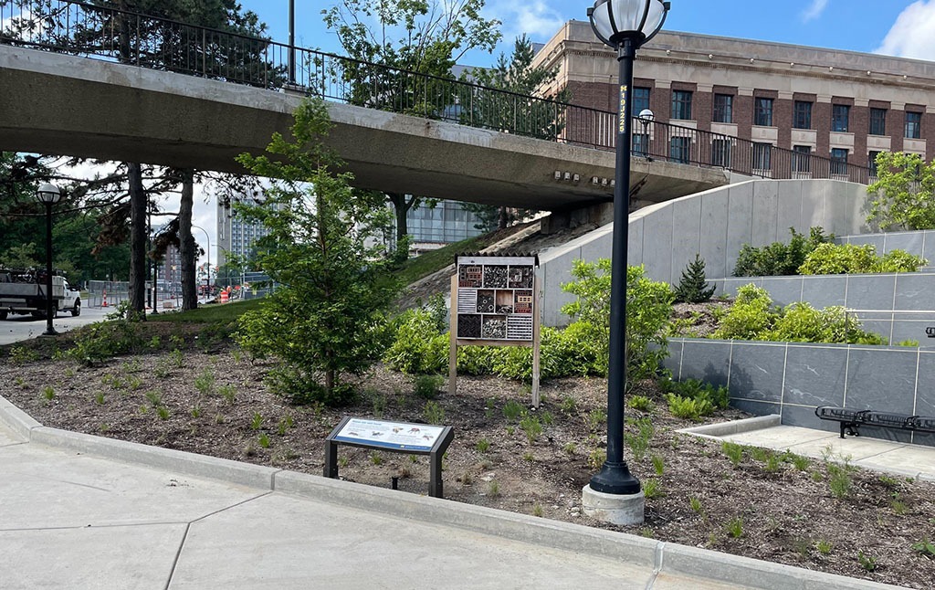 An outdoor area with a raised concrete walkway supported by a metal railing. In the foreground, a landscaped area with small plants and bushes is bordered by a paved sidewalk. Two informational signs are prominently displayed within the landscaping; one is mounted on a stand and visible in the center, while the other is closer to the ground near the walkway. A streetlight with a round lamp is also present, and modern buildings are visible in the background. The sky is partly cloudy with blue patches.