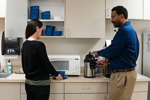 A woman putting away dishes and a man pouring himself coffee talk to each other in a kitchen.