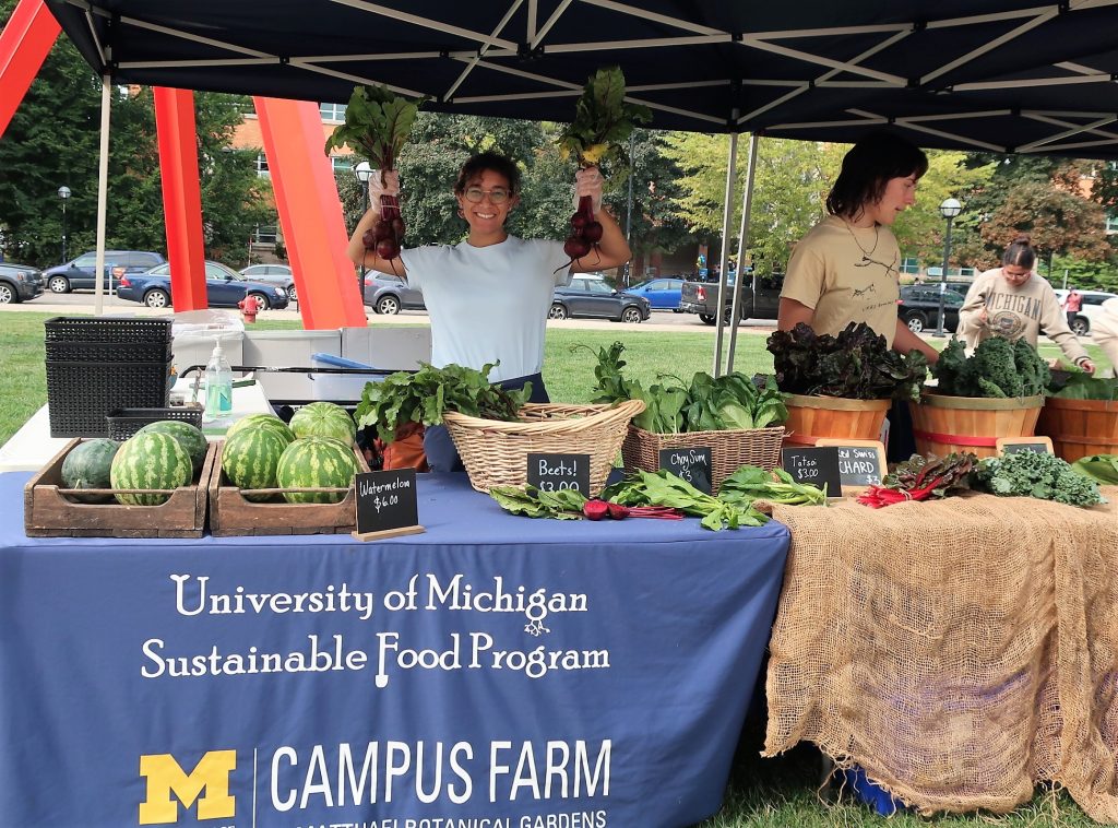 a young woman holds up fresh beets and grins at a farmers market stall