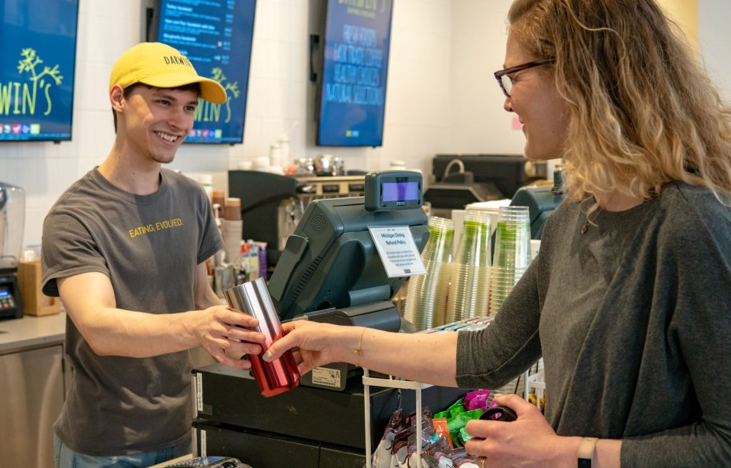 a man working behind the counter of a coffee shop accepts a reusable mug from a customer