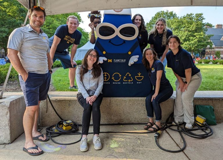 a group of people smile while standing around a mascot costume that has sunglasses, a smile, and overalls with icons about waste prevention