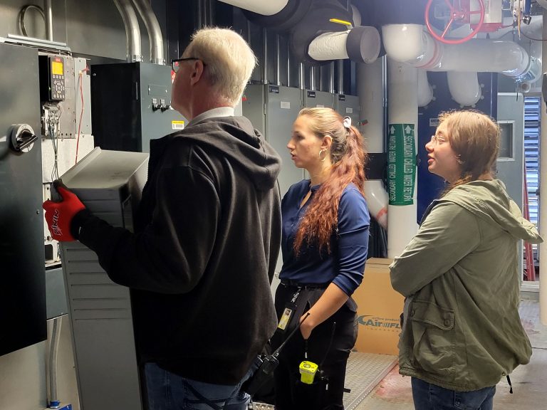 Two OCS interns learn about temperature controls from a technician in a maintenance room.